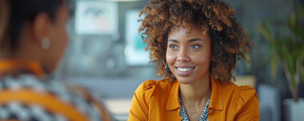 Woman with curly hair smiling at another person in a bright room.