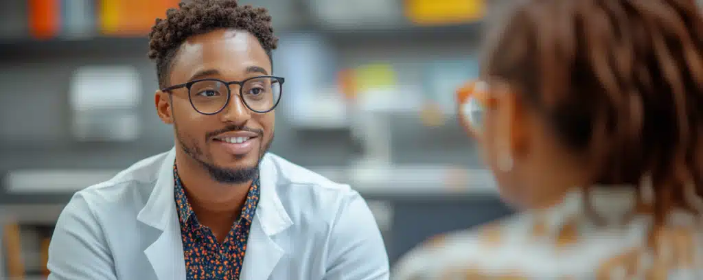 A health psychologist in glasses and a lab coat smiles while talking to another person in an office setting.