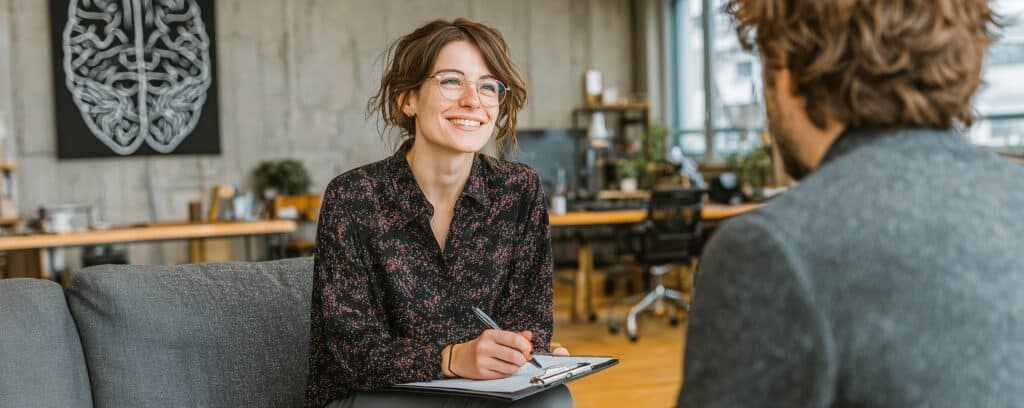 Smiling woman with glasses holds a clipboard, talking with a man in a modern office setting.