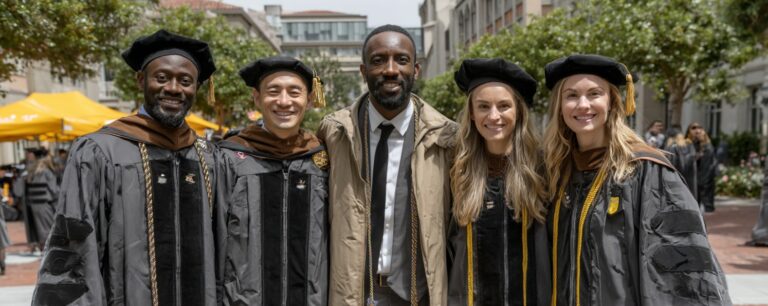 Five people, including four in graduation robes and one in a suit, smile together outdoors at a graduation ceremony.