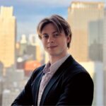 Young man, IO Psychologist Joe Kaul, stands in a blazer with arms crossed, smiling, with a city skyline blurred in the background.