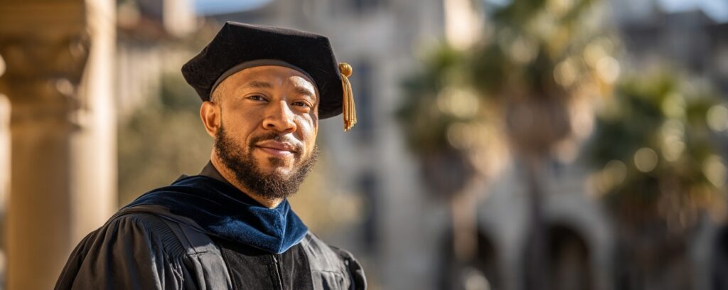 Man in academic regalia and graduation cap standing outdoors, smiling, with blurred buildings in the background.